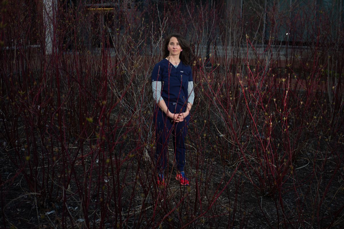 Nurse in blue scrubs stands in a dark field with dark foliage