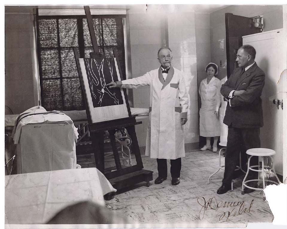 Black and white photo of a doctor in a white lab coat pointing to an illustration on an easel, while a nurse and man in suit look on