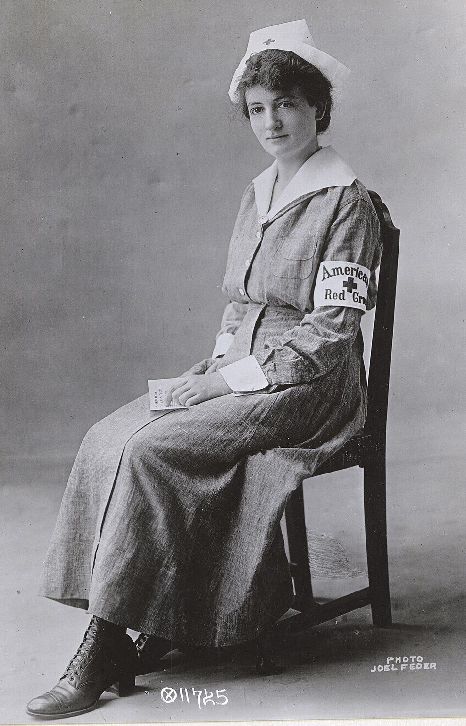 Black and white photo of a seated woman in nursing uniform: white hat with small cross, buttoned up dress with white cuffs and collar, and a white armband with a cross and the words "American Red Cross"