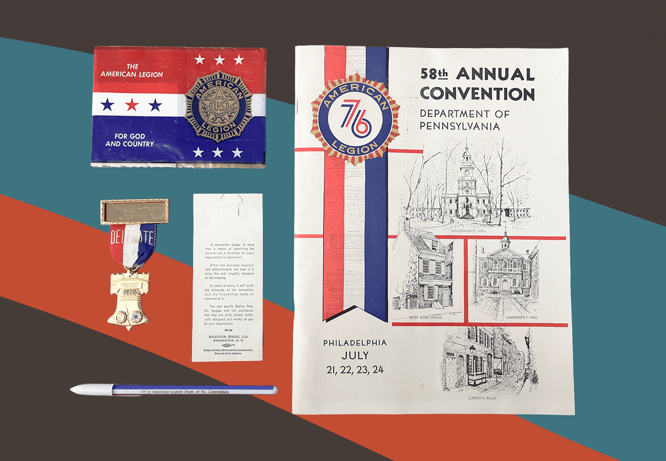 Photo of an assortment of red, white, and blue collateral from the 58th Annual Convention of the American Legion. Items include a program booklet, pin shaped like the Liberty Bell, and pen. They sit against a brown background with a muted red and blue swoosh running through the middle.
