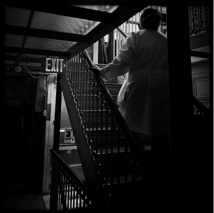 A black and white photo of a woman in a lab coat ascending an antique staircase