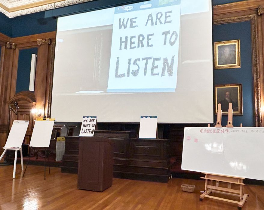 White boards lined up under a projector screen displaying the message "We are hear to listen"
