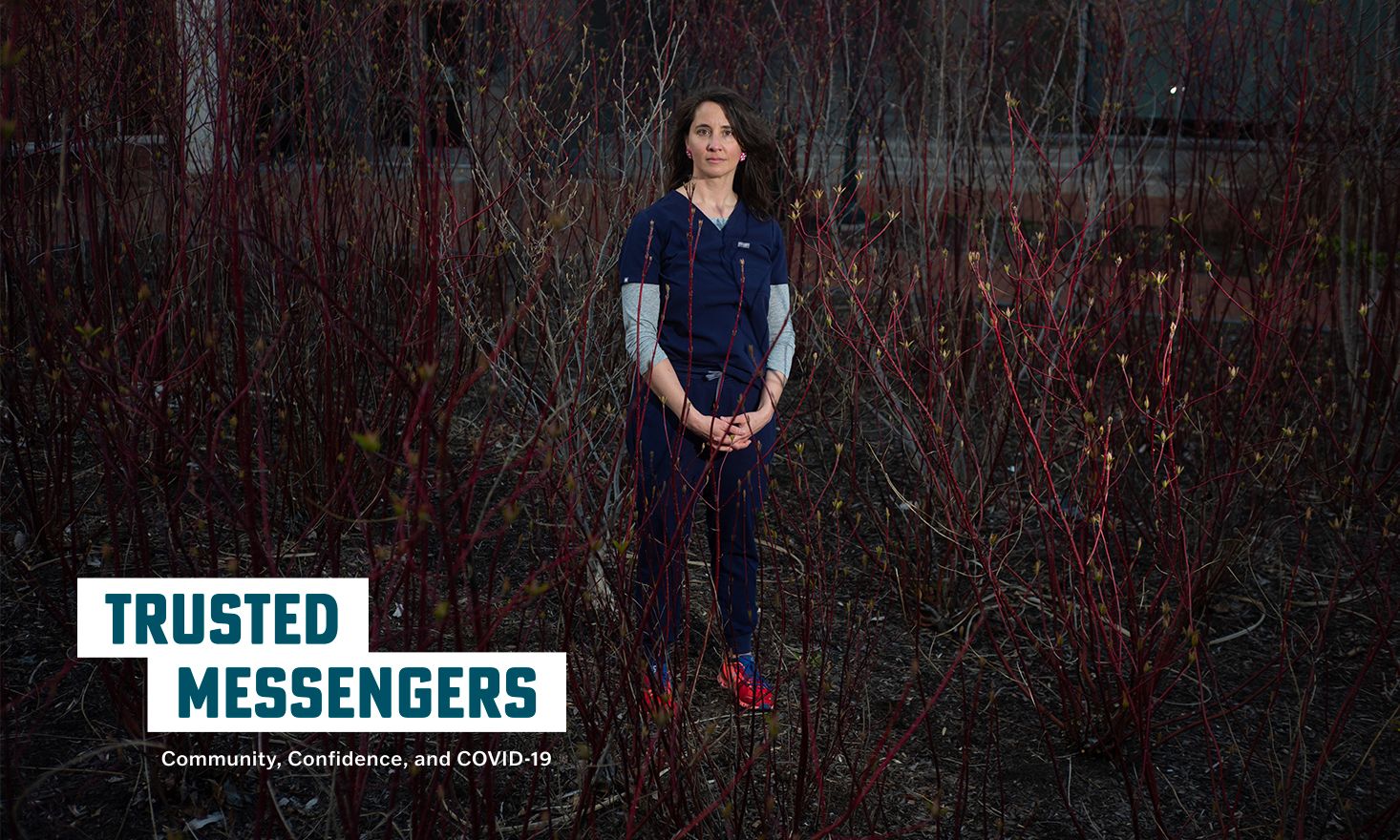 Nurse in blue scrubs stands in a dark field with dark foliage, with the text "TRUSTED MESSENGERS"