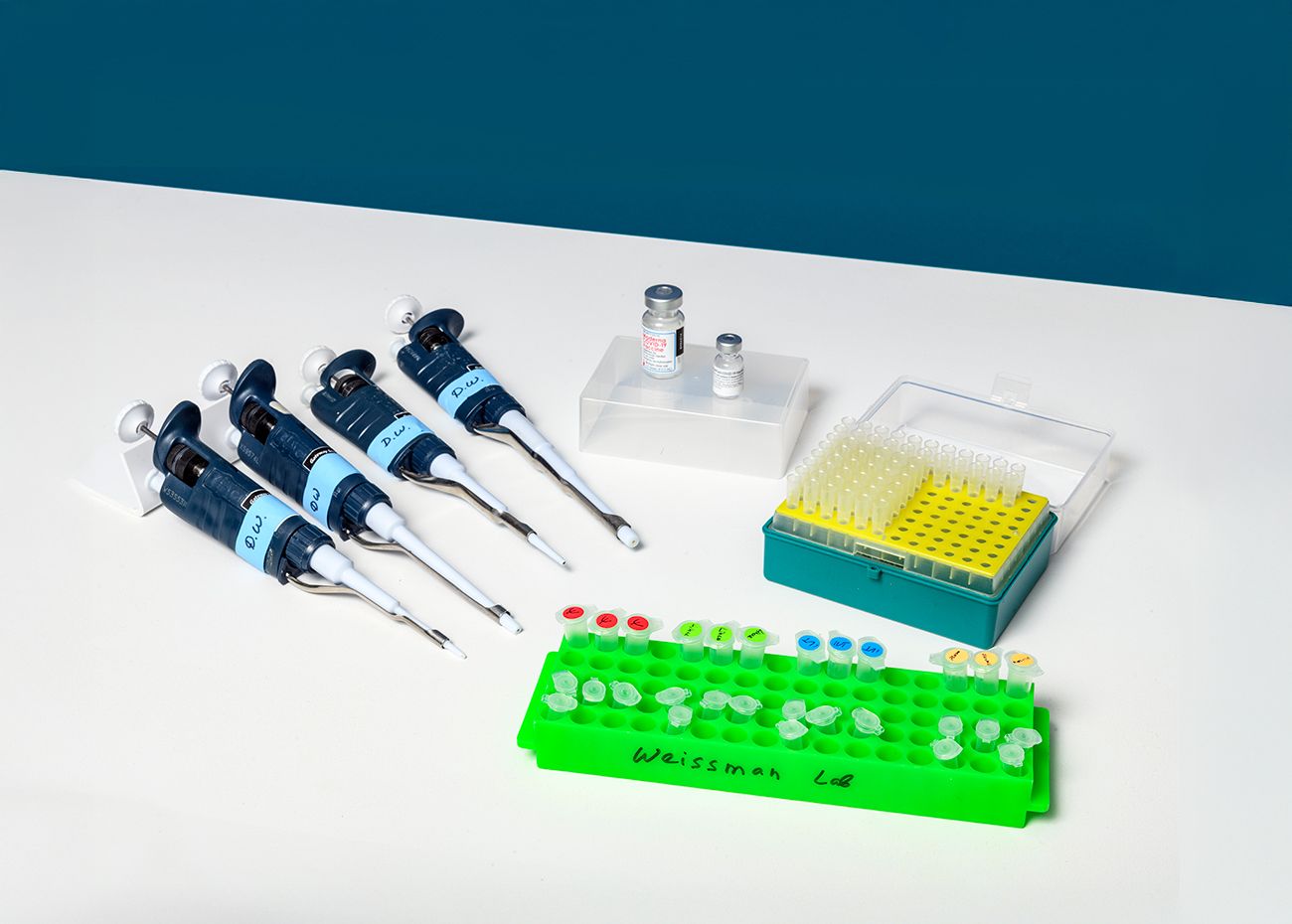 Assortment of pipettes, vials, and small tubes, with a bright green tray labeled "WEISSMAN LAB" on a white surface against a blue background