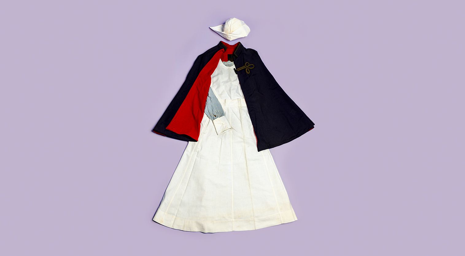 Nursing uniform consisting of a white hat, blue striped shirt, white apron, and navy cape with red lining, against a light purple background