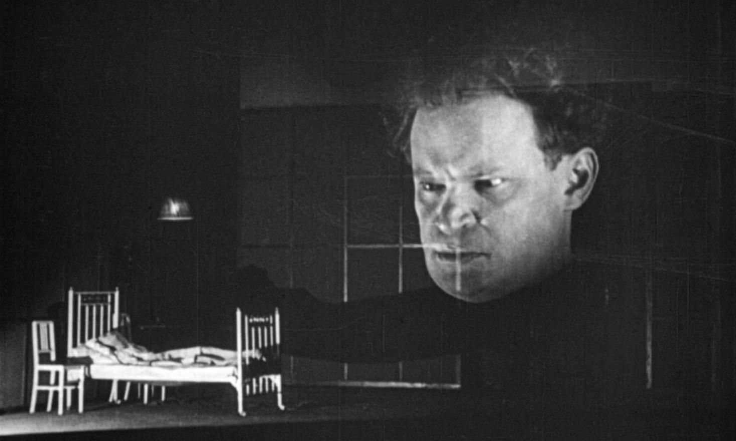 Old Black and white film still with single bed and chair in lower corner and large ominous face looming over