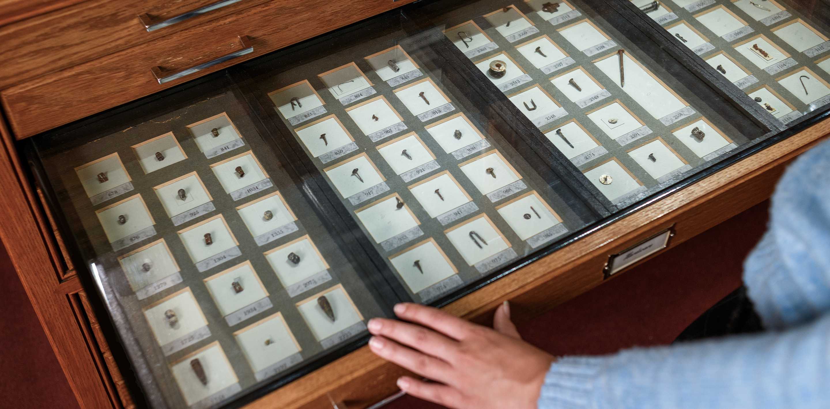 A person in a light blue sweater rests their hand on an open drawer holding dozens of tiny objects from the Chevalier Jackson collection