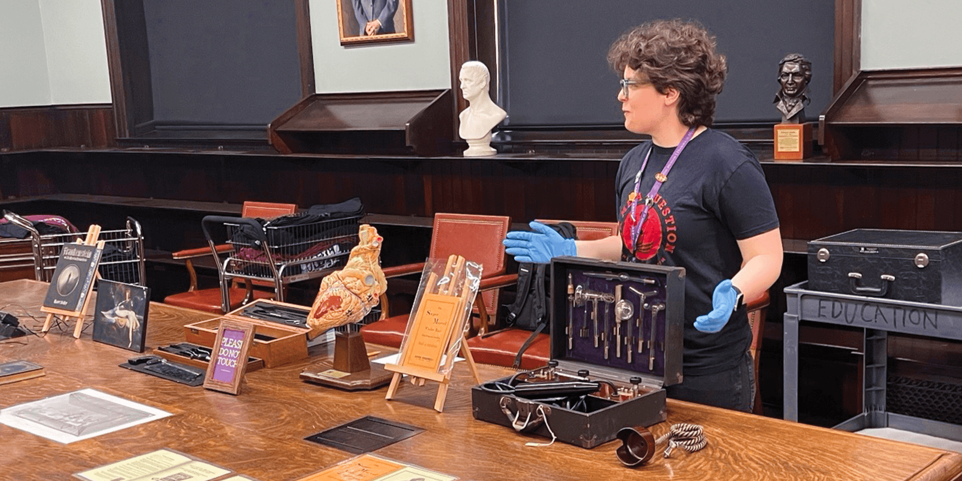 A Museum docent with short brown hair, a black tshirt, and blue gloves stands in front of a table where various Museum collections items are laid out, including a model of the human heart