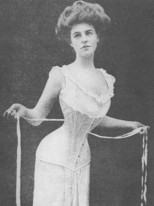 A young white woman. She wears a frilly, sleeveless dress with a corset along her midsection. She holds tailor's tape along a very thin waist.